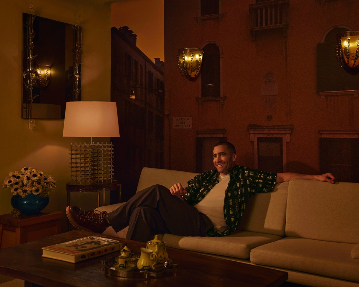 Jake Gyllenhaal sitting on a beige sofa in a dark-toned room, with brown wooden features.