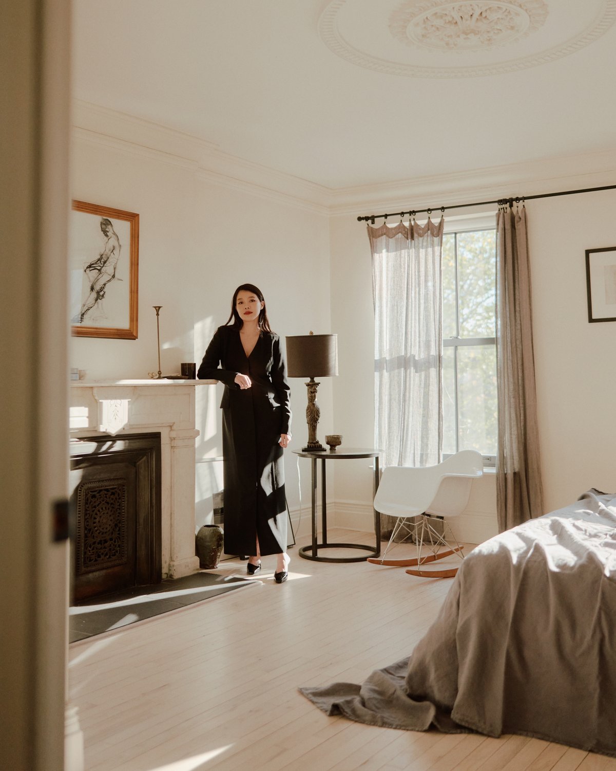 Ein sanft beleuchtetes Schlafzimmer mit Kamin, hellen Holzböden, leichten Vorhängen und einem Bett in neutralen Tönen, eingerichtet mit einem Loungesessel und minimaler Dekoration.