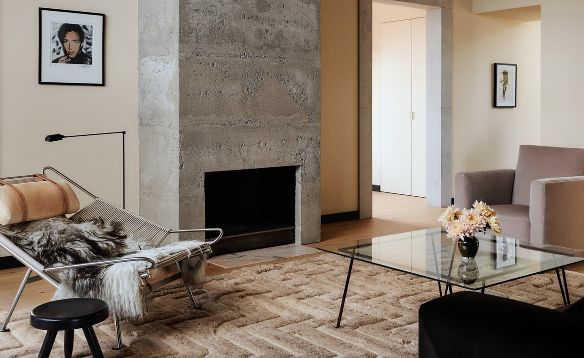 Der Teppich Boho 02 – Taupe Mix in einem warmen Wohnzimmer mit Betonofen und modernen Möbeln. Gestylt mit einem Glastisch, einem Loungesessel und neutralen Farbtönen.