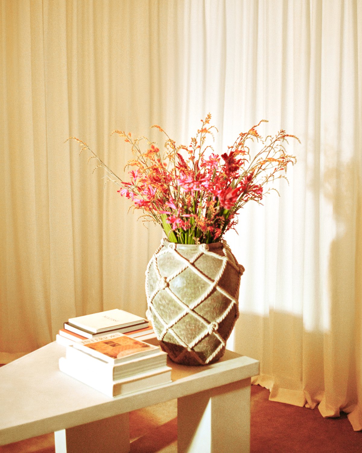 The Grand curtains in Perfect White depicted behind a large ceramic flower vase on top of a white, triangular office table next resting on plush wool rug Grand in Leo in the Stockholm Flagship store upstairs curtain room.