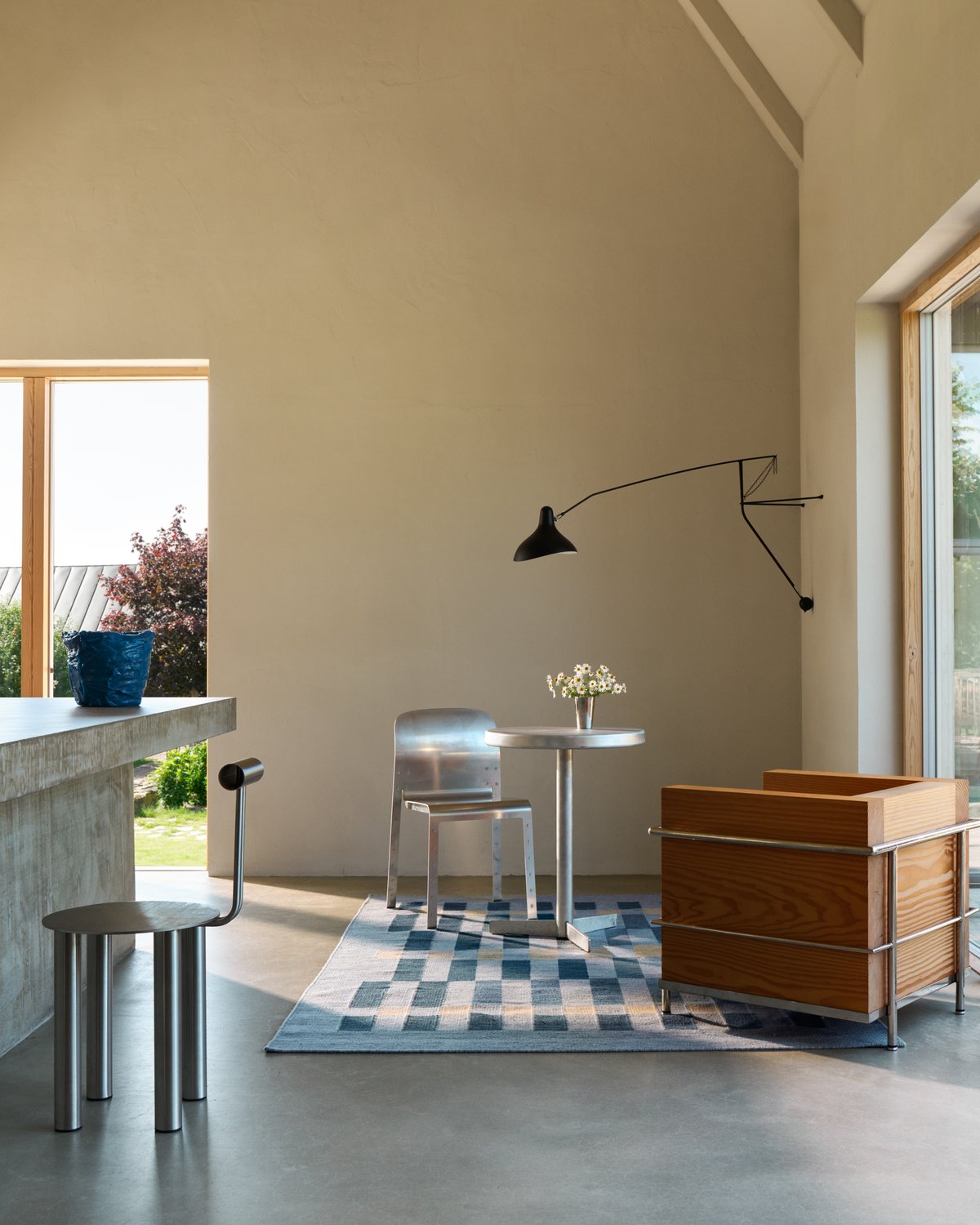 A lounge area with Tradition 01 in Blue, where the rug sits under a small steel table and chair, next to a square wooden armchair and a black wall lamp.