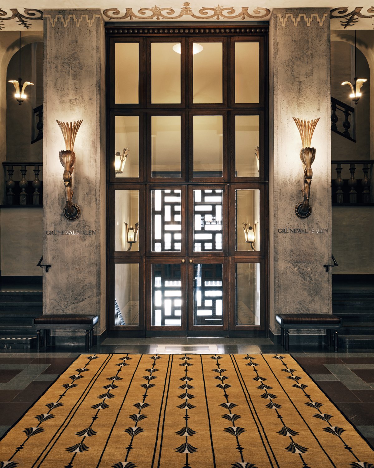 Grace 04 – Camel — displayed in the grand hall of Stockholm Concert Hall. The camel rug features repeating black tassel-like motifs arranged in symmetrical vertical lines.
