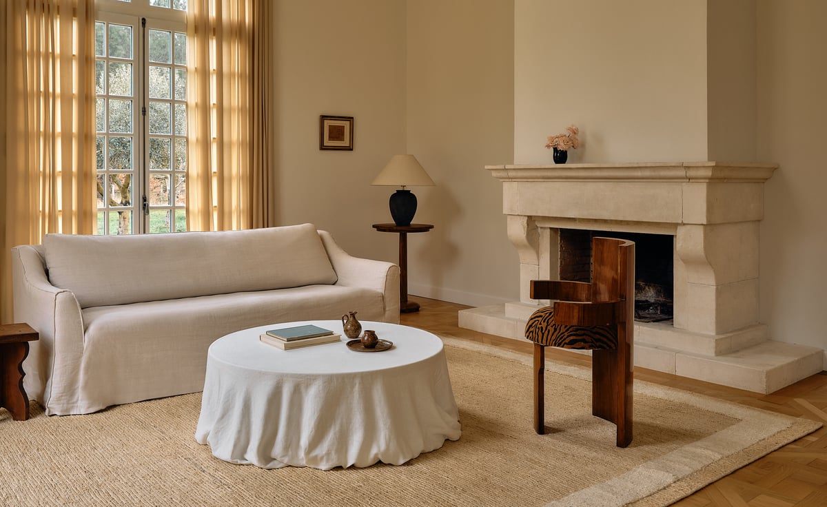 The Jute Frame rug in Cream is shown in a bright living room with a cream sofa, round draped coffee table, and stone fireplace, creating a natural and elegant atmosphere.