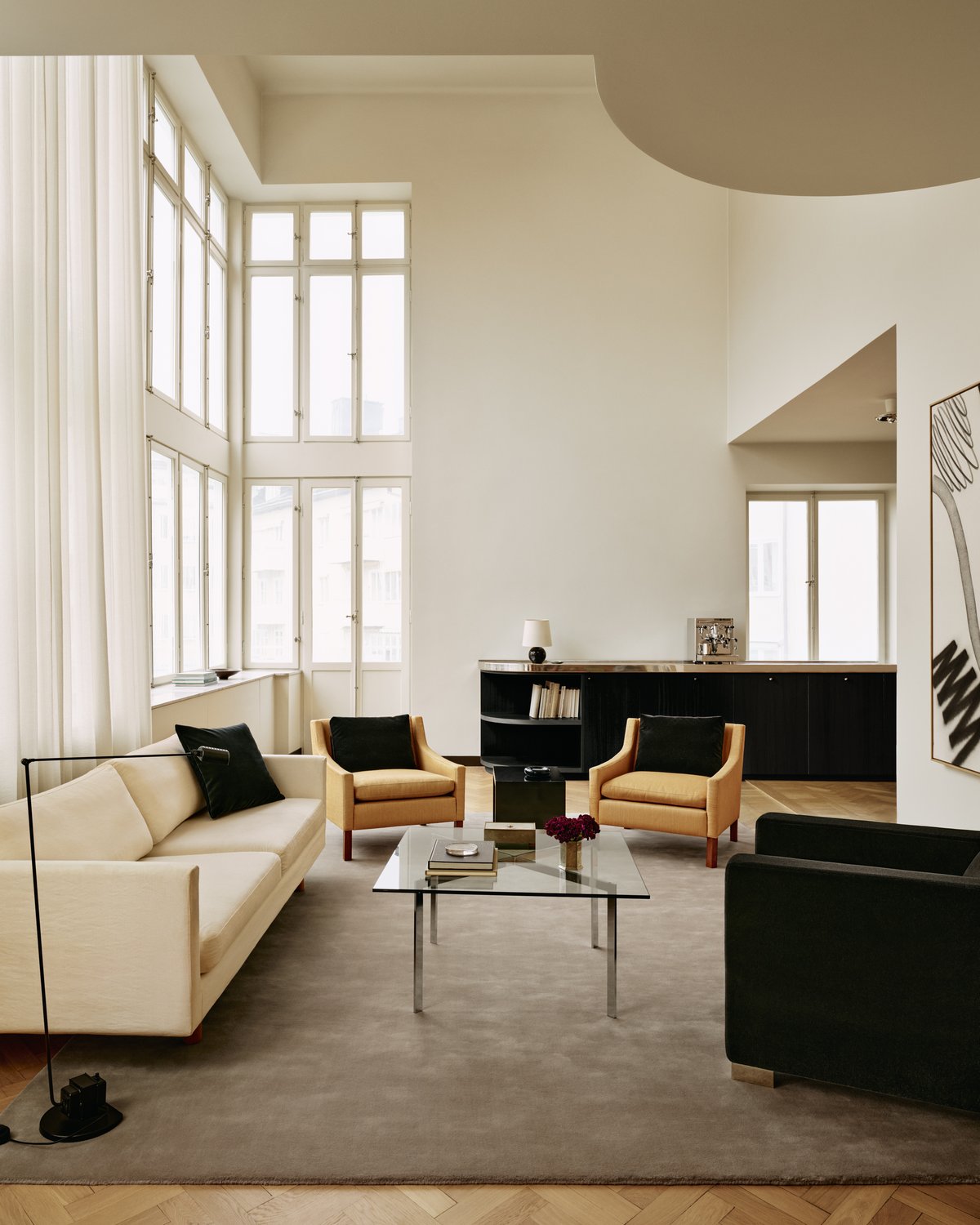 Tapis Grand en ton neutre Warm Gray dans un salon baigné de soleil, associé à un canapé crème, des chaises noires et marron foncé, ainsi qu’une table basse en verre avec une structure métallique.