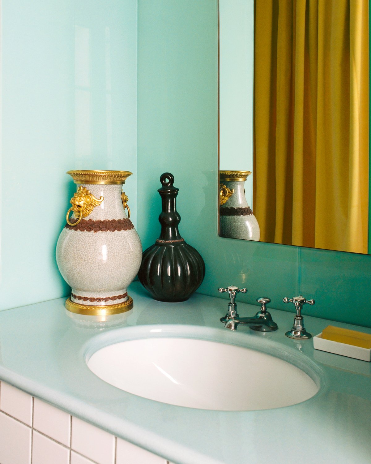Meuble de salle de bain avec lavabo bleu pâle et miroir, mis en scène avec des vases décoratifs, avec les rideaux The Grand en Leo reflétés en arrière-plan.
