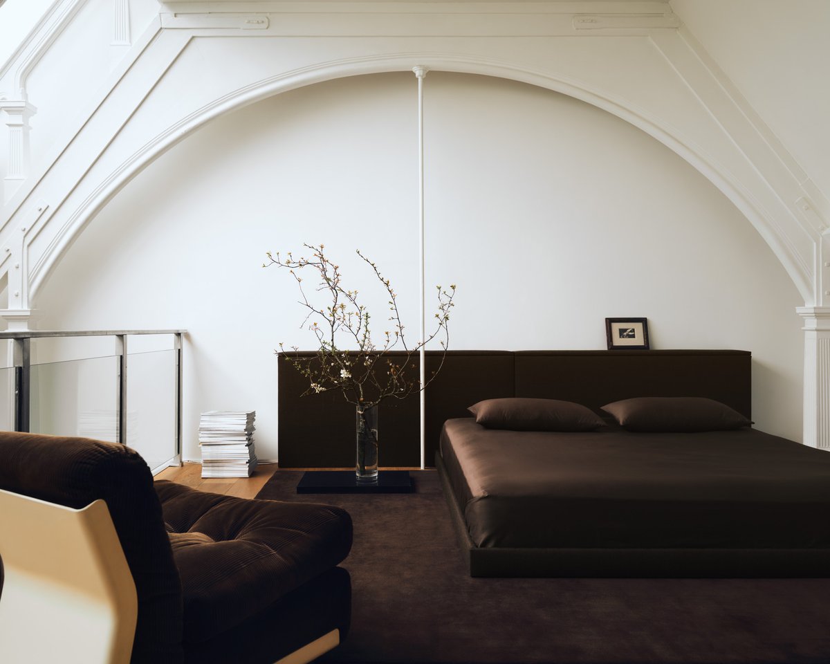 Der Teppich The Grand in Walnut, inszeniert in einem Loft-Schlafzimmer in Mailand mit einem niedrigen dunklen Bett unter einer weißen Gewölbedecke, kombiniert mit skulpturalen Sitzmöbeln und minimalem Dekor für eine warme, architektonische Atmosphäre.