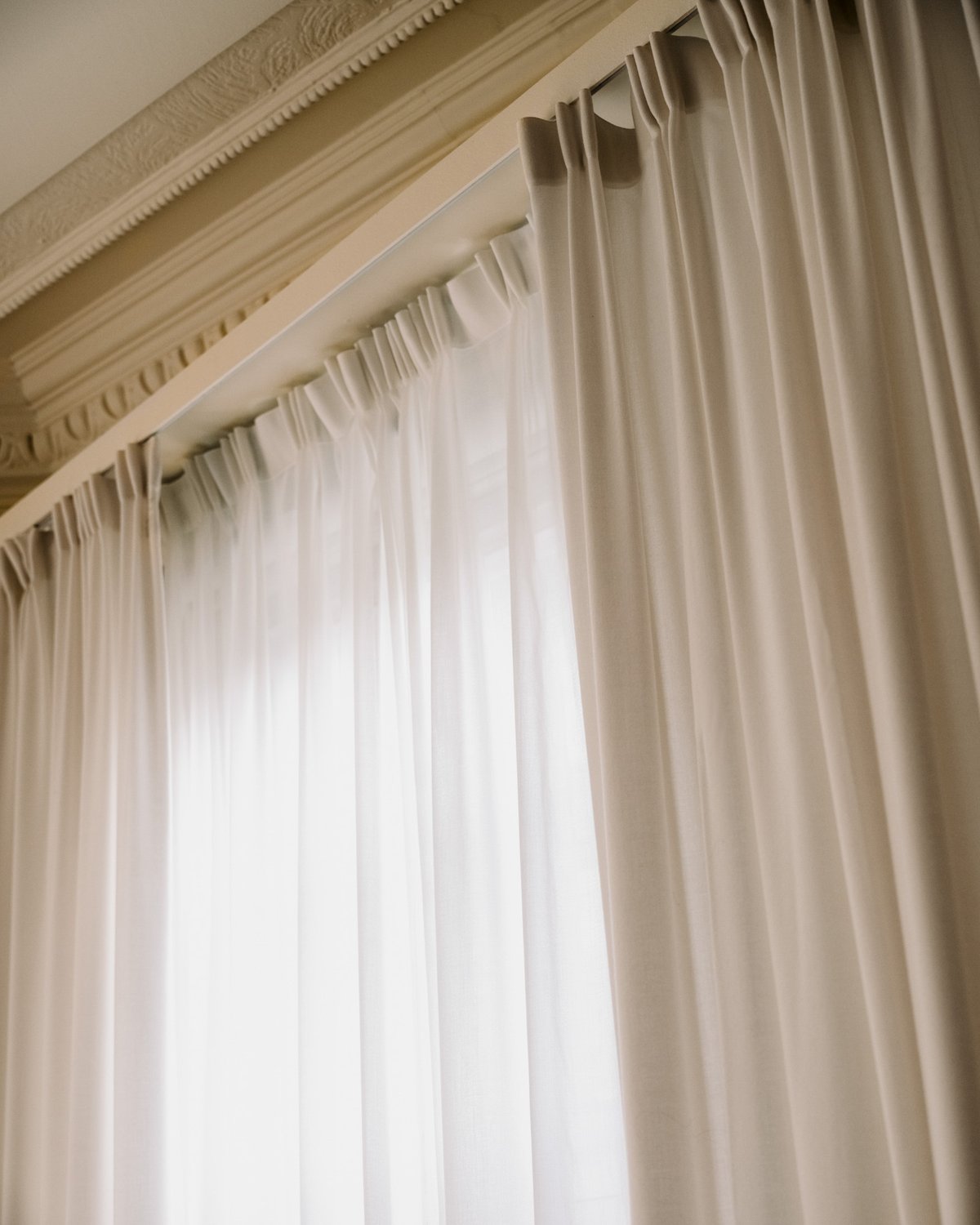 The Grand curtain in Perfect White shown by a window, displaying a soft white fabric with a smooth woven texture and full-length vertical folds filtering light.