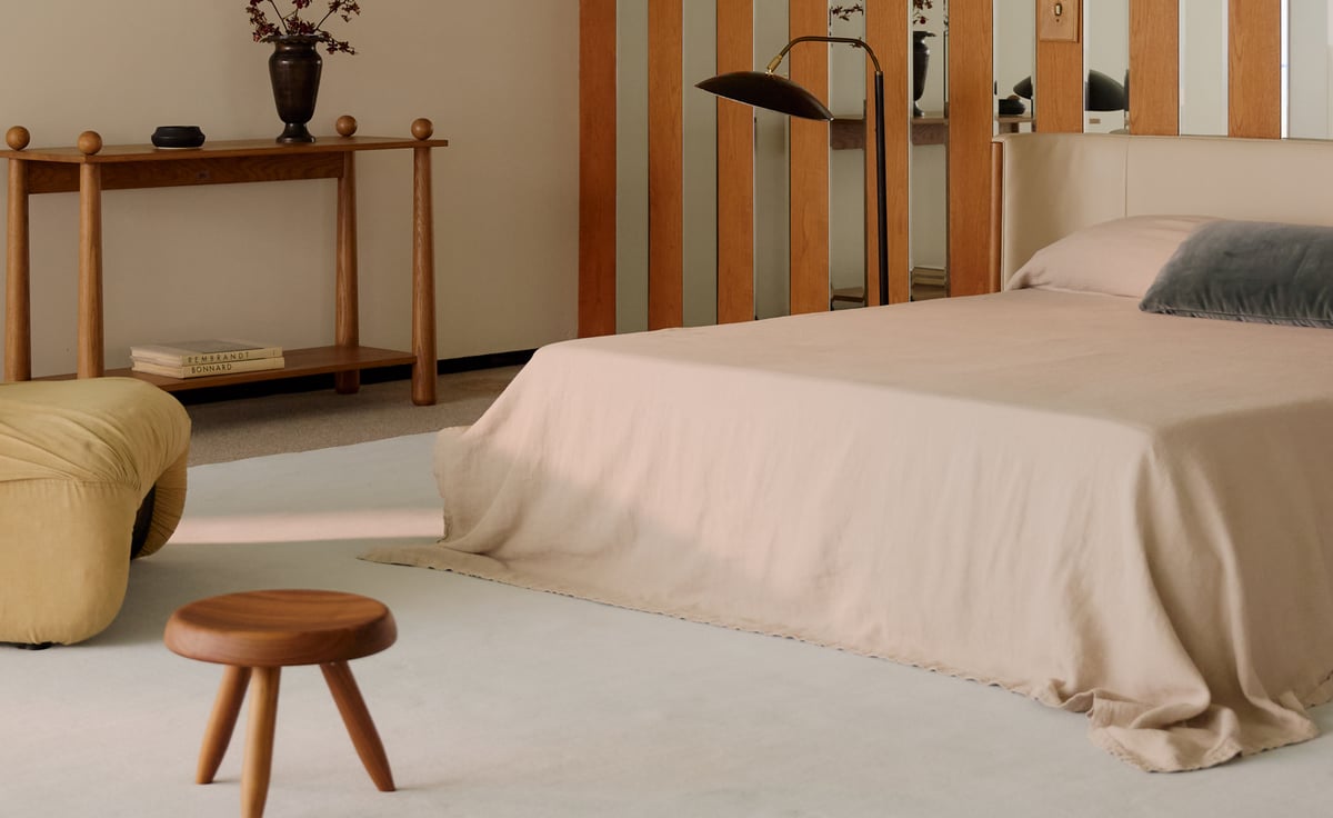 The Grand rug in Powder Blue is shown in a minimal bedroom with soft neutral tones, a low wooden stool, and sculptural details, creating a calm and refined atmosphere.