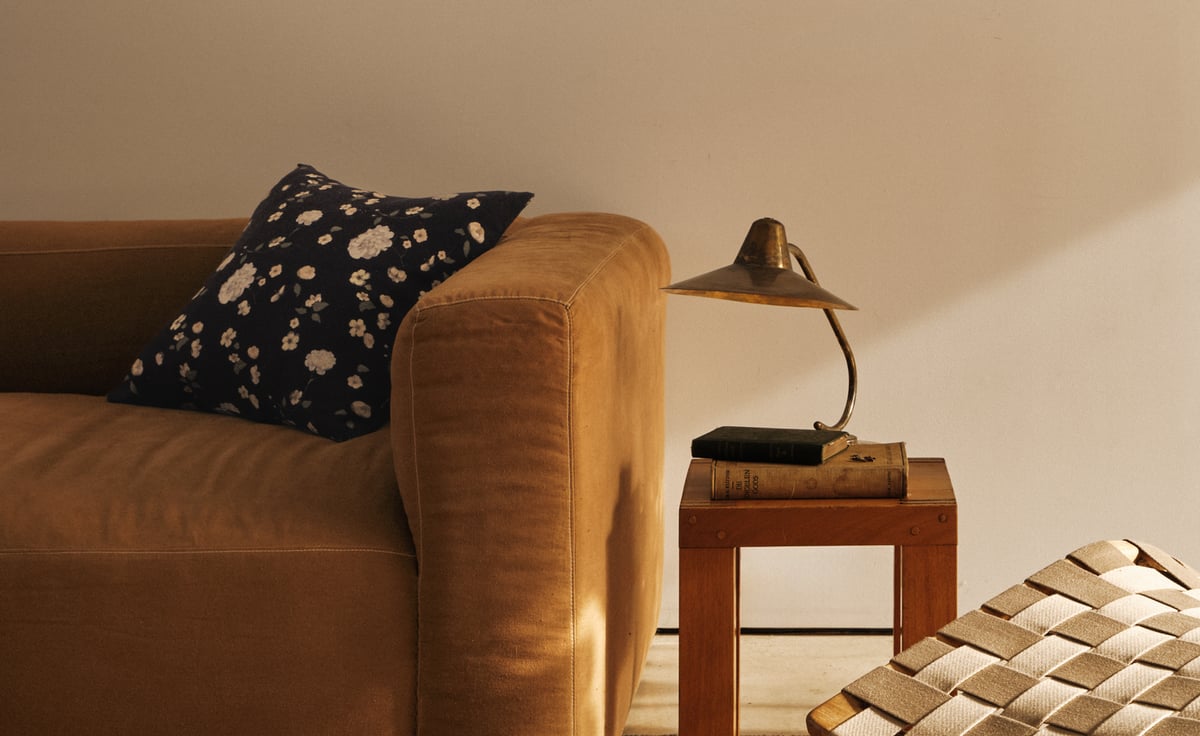 A sofa upholstered in Studio Linen in Brandy, styled with the Honey Rose – Midnight cushion. A small wooden side table stands beside it with a lamp and a few stacked books.