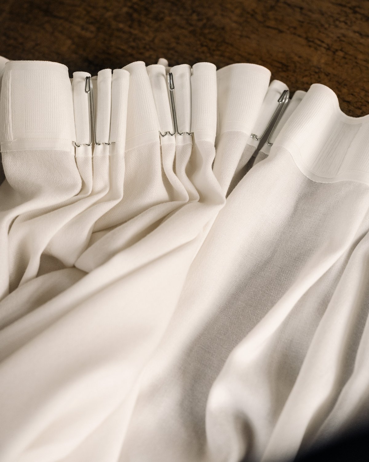 Close-up of The Grand curtain in Perfect White, showing a soft white fabric with a smooth woven texture, gathered header, and metal hooks forming structured pleats.