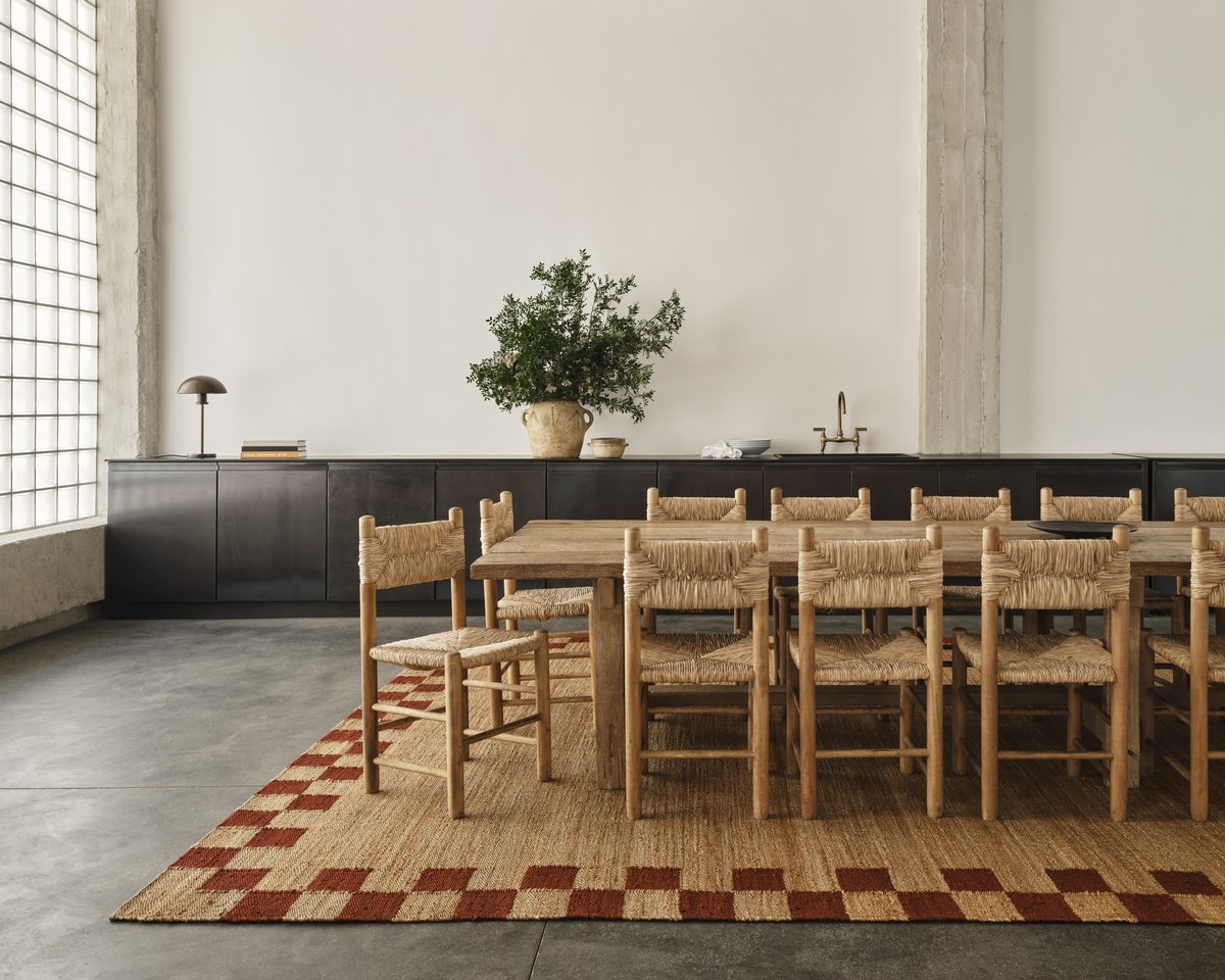 A big Jute Border rug in Red displayed under a huge dining table with multiple wooden and rattan chairs around it. The space is large, bright with a dark kitchen in the back.