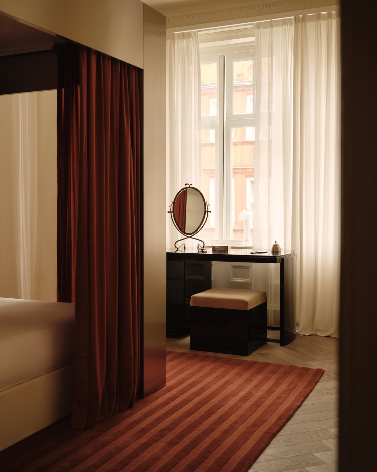 The Park 03 rug in Burgundy is shown in the Nordic Knots store on Mount Street in London, styled in a softly lit bedroom with Burnt Red curtains, light sheer drapes, and a black vanity table, creating a warm and elegant atmosphere.